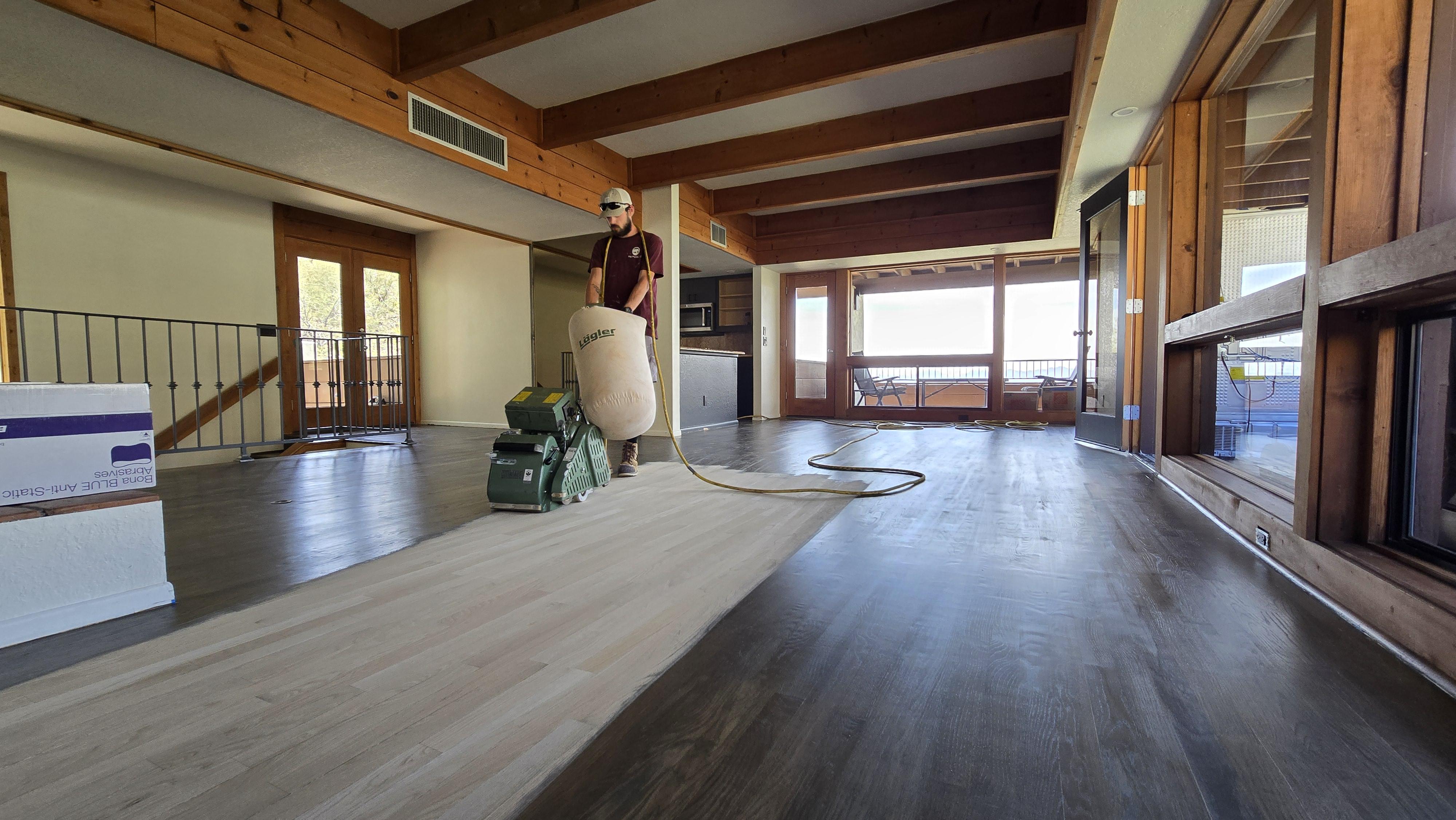 A professional contractor operating a green Hummel floor sanding machine on light-colored hardwood planks inside a residential room with natural light and an arched doorway.