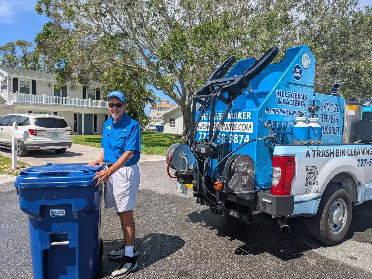 A man in a blue shirt standing next to a trash bin and a blue trash bin cleaning truck.