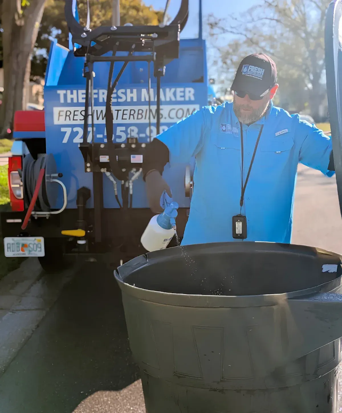A man in a blue shirt and sunglasses sprays cleaning solution into an empty trash bin next to a Fresh Maker truck.