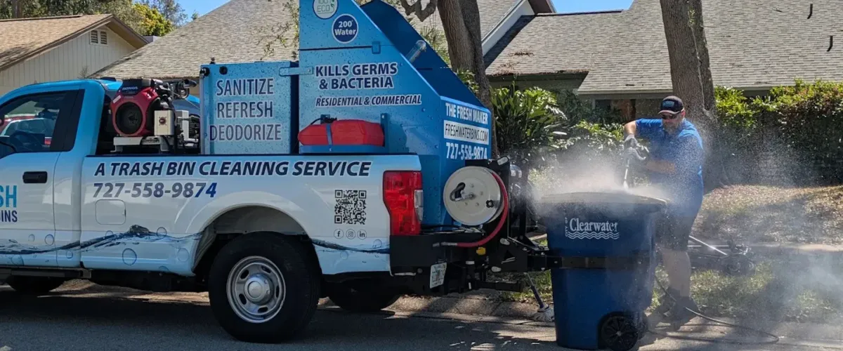A man in a blue shirt uses a power washer to clean a blue trash can next to a white service truck.