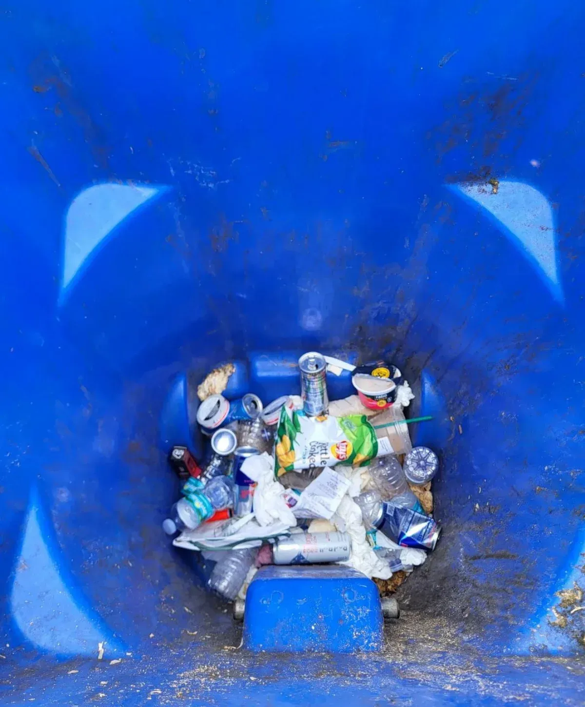 A top-down view inside a blue trash can overflowing with assorted garbage, including cans, plastic bottles, and wrappers.