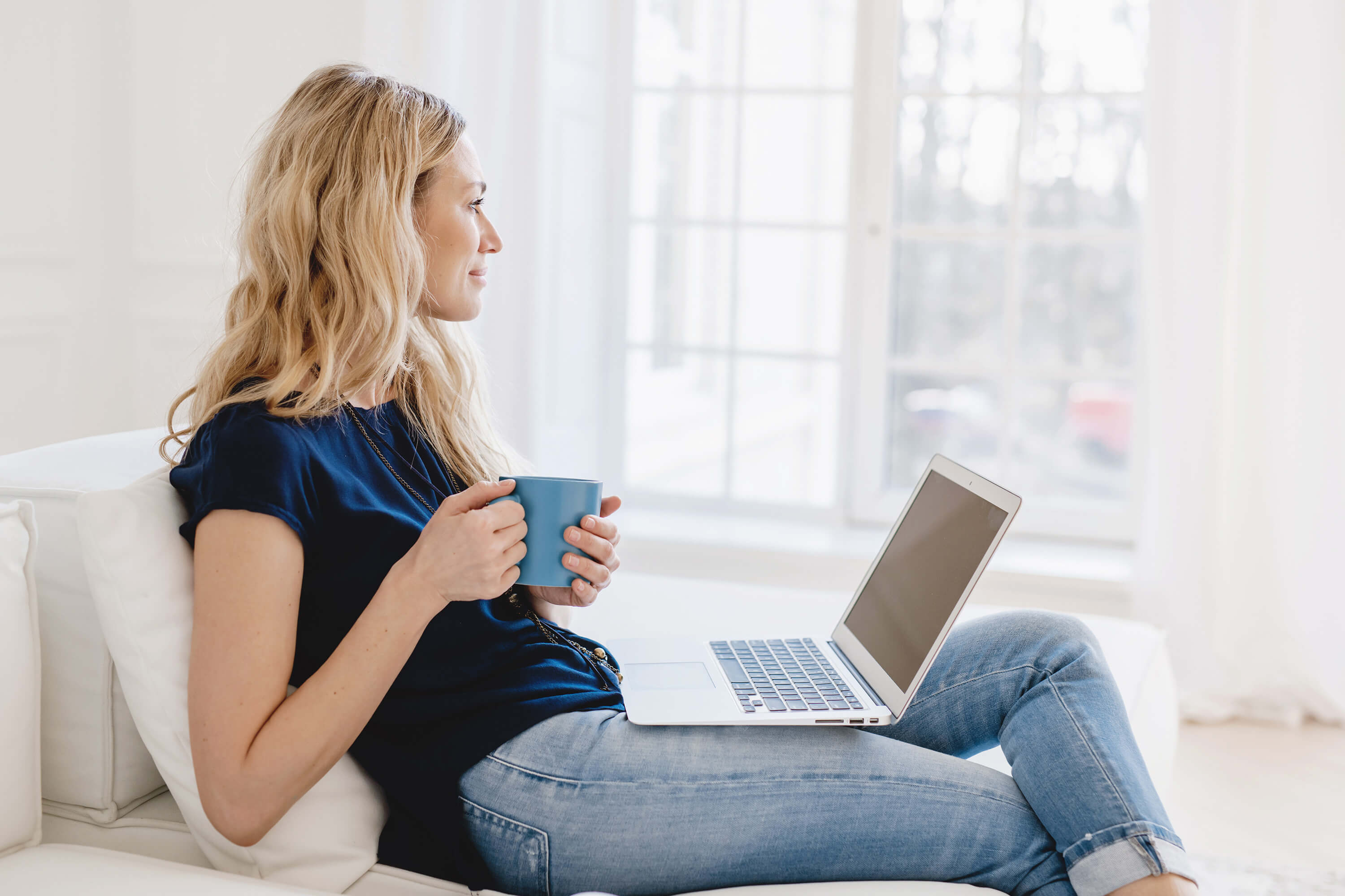 Entrepreneurial woman in jeans and a dark blue t-shirt holding a coffee cup, with laptop on her lap, staring off into the distance wondering how she can get more strategic support in her business