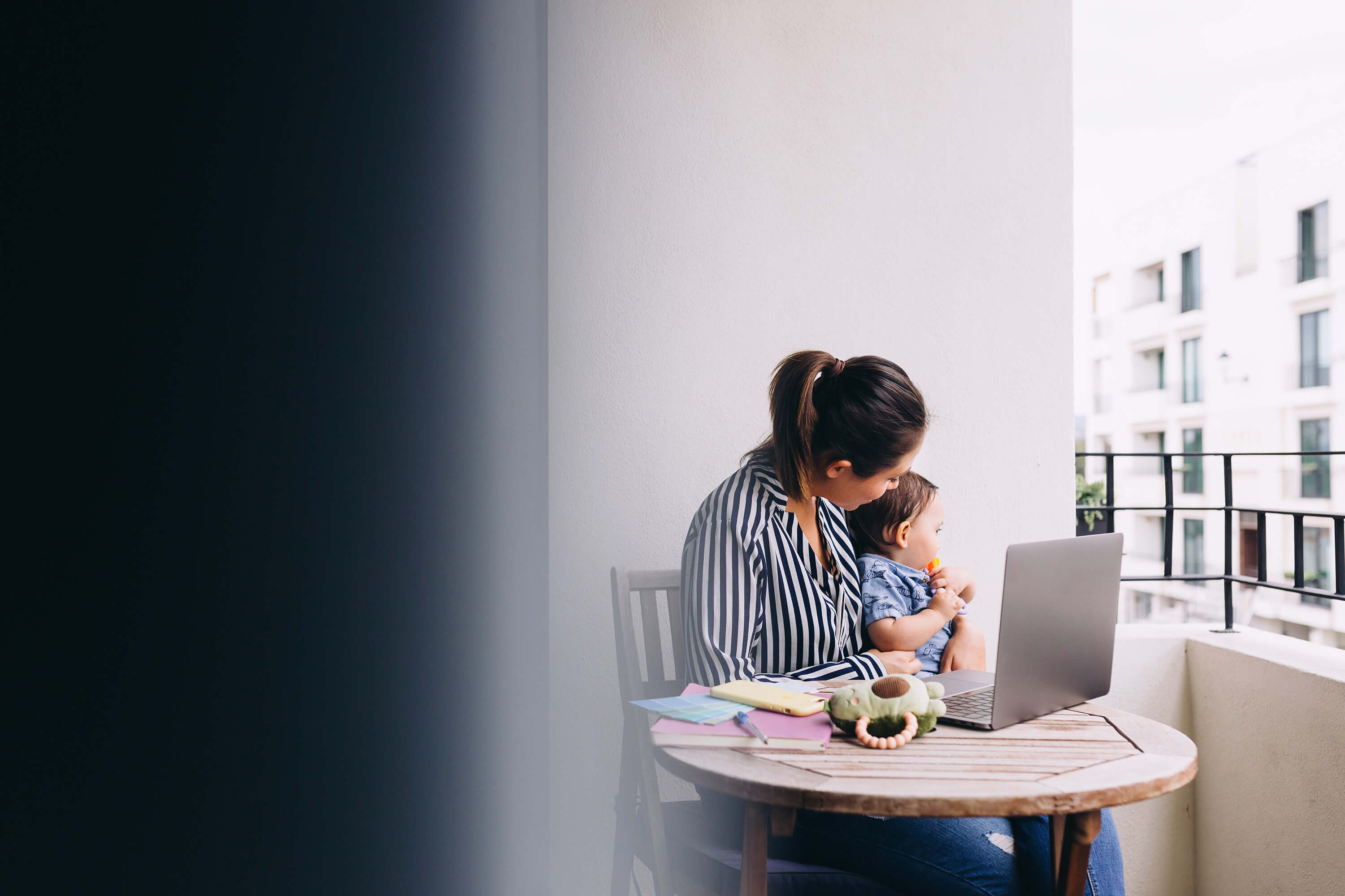 Intuitive business woman sitting at a table on a balcony, working with a laptop in front of her and a small child on her lap