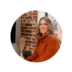 Headshot of Jessamyn Anderson, founder of Root to Rise Creative, wearing a burnt orange sweater, sitting in front of a painting hung on a brick wall