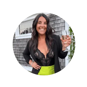 Headshot of Julie Costa, leaning against an external house wall with gray shingles, wearing a black bustiere and a ime green skirt, holding a glass of champagne