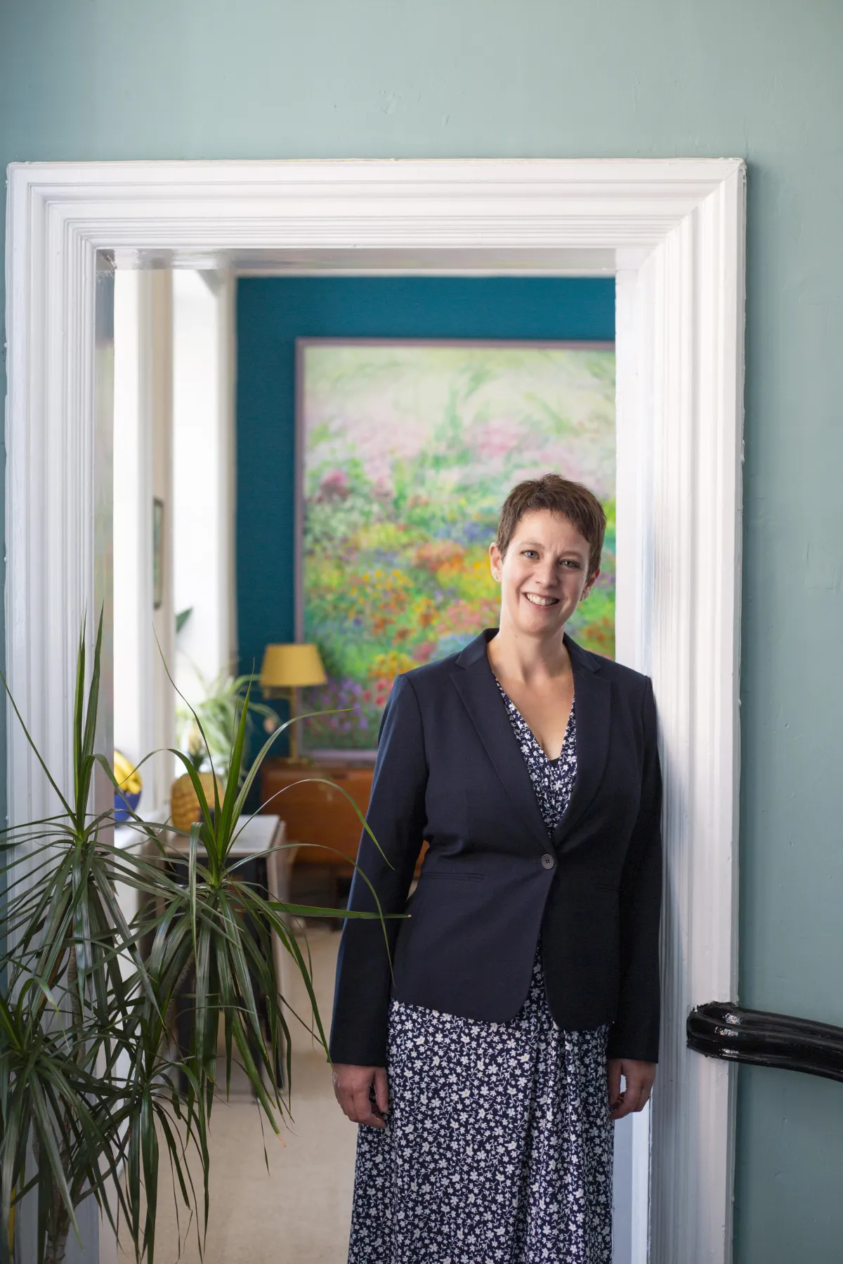 Picture of Charlotte wearing a blue dress and blazer, leaning against a door frame and smiling