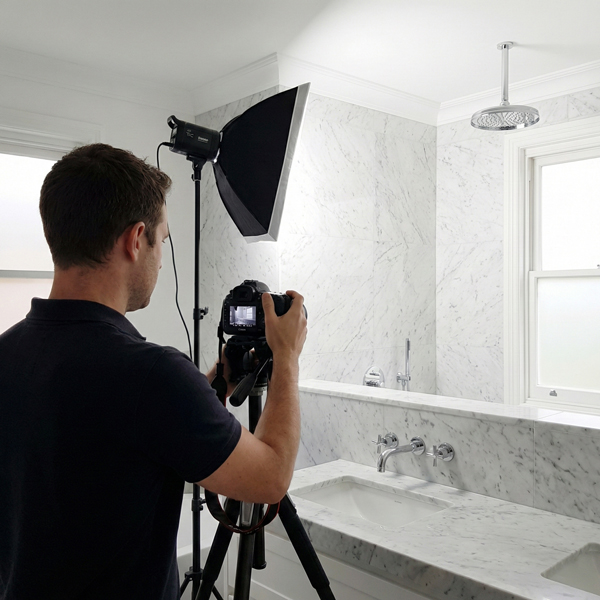 Professional photographer capturing interior photos of a modern bathroom featuring marble finishes and chrome fixtures, using a camera with softbox lighting and flash for property photography.