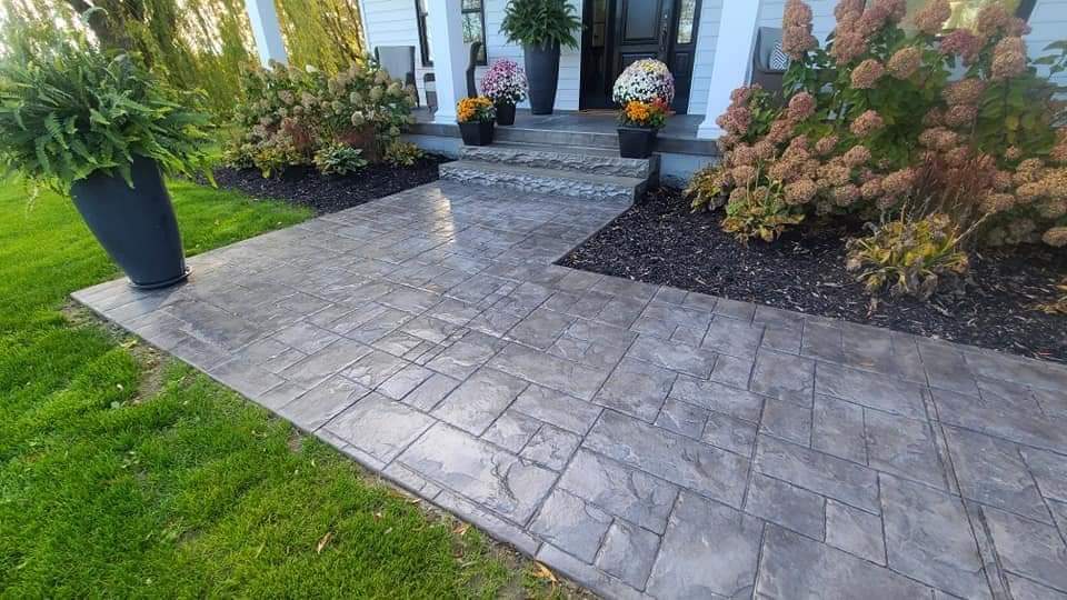 A close-up of a stamped concrete walkway with a natural stone pattern, bordered by vibrant flower beds. The surface is clean and detailed, showing the precision of the stamping and the integration with the surrounding landscape.