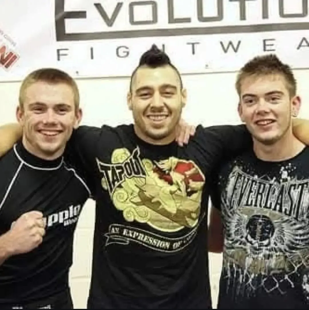 Two men posing in Muay Thai stances outside a gym, with a fighters-in-the-ring graphic and class times in the window behind them.
