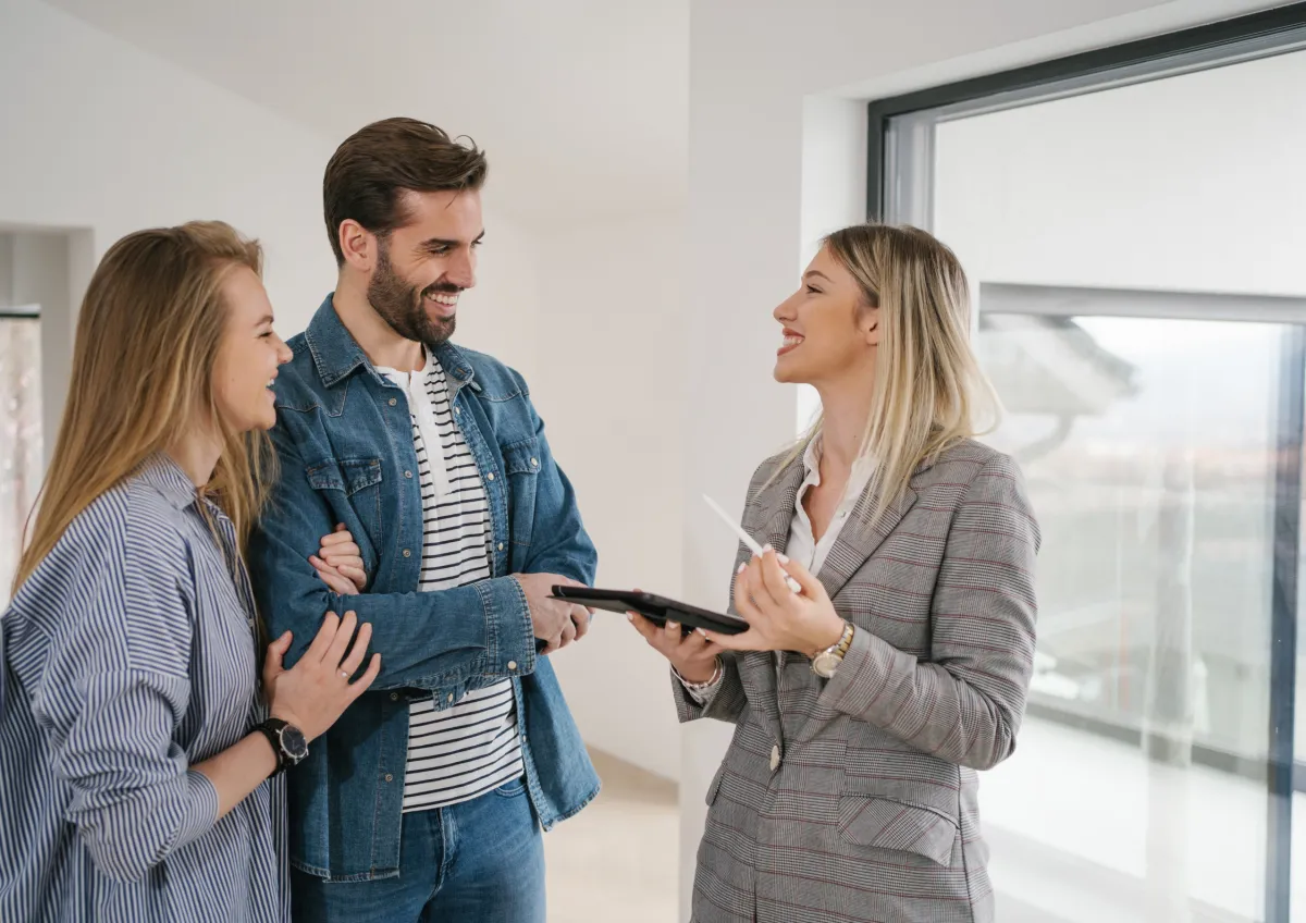 A real estate agent in a smart-casual outfit making a friendly phone call from a bright, modern home office, with a laptop open and a notepad filled with client names. The agent is smiling, focused, and appears to be re-engaging with a past client.