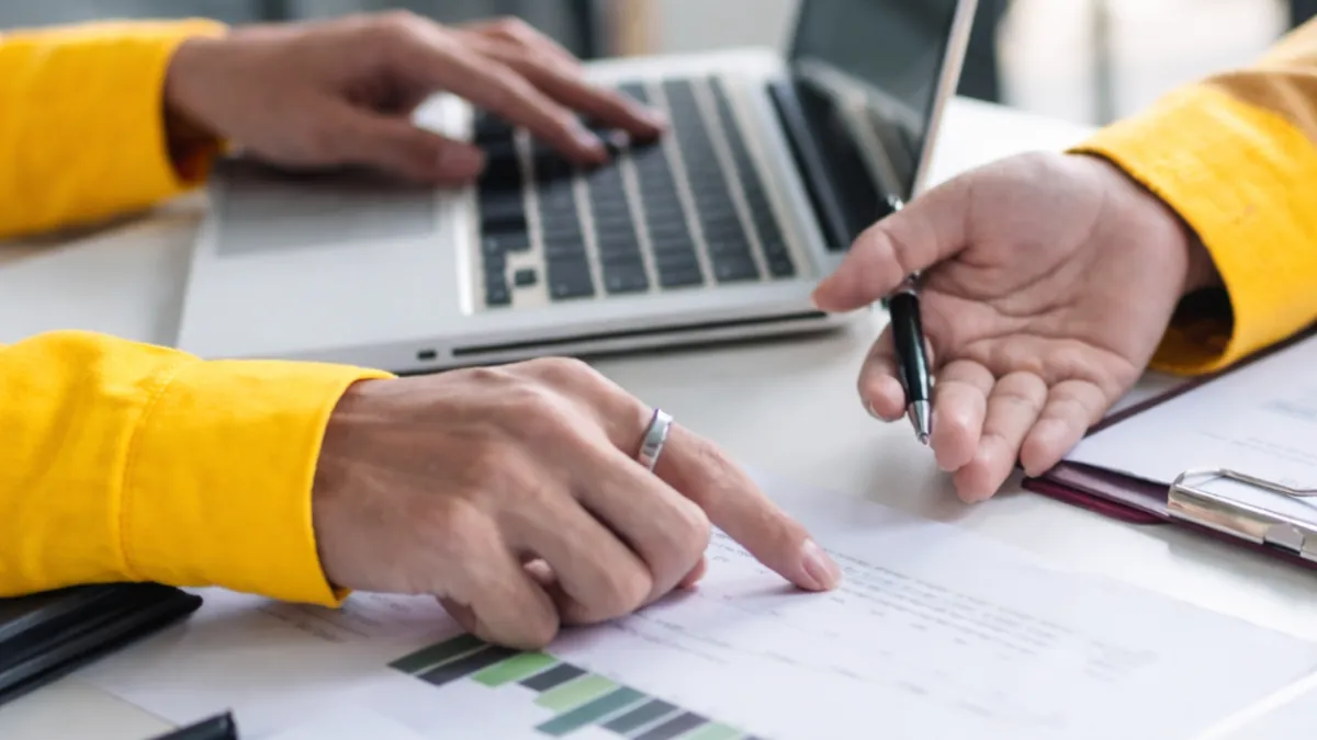A real estate agent in a smart-casual outfit making a friendly phone call from a bright, modern home office, with a laptop open and a notepad filled with client names. The agent is smiling, focused, and appears to be re-engaging with a past client.