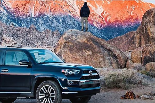 Toyota SUV parked in front of mountains with man standing on a boulder