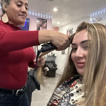 A professional stylist with short, vibrant hair is applying color to a client's hair in a chic, sunlit salon. The client, a young woman with medium-length hair, smiles in the mirror. The salon features modern decor with gold accents and lush plants, creating a lively, welcoming atmosphere.