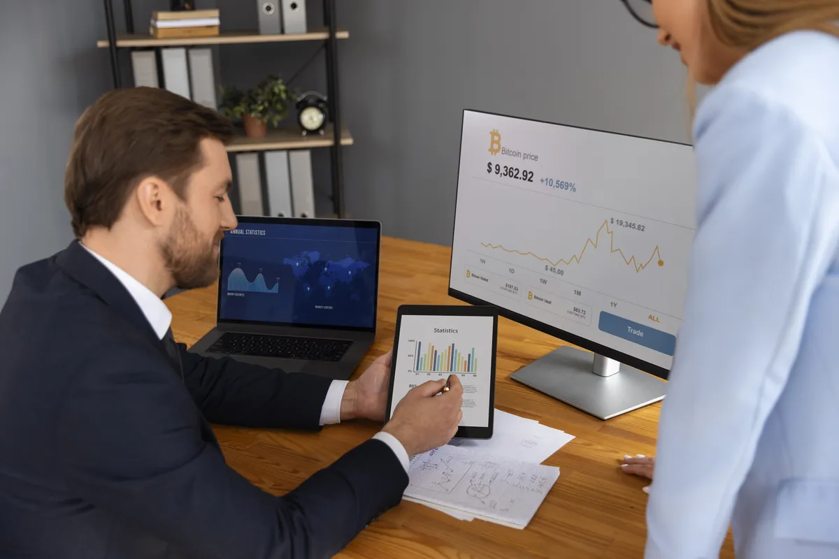 Two professionals, a man and a woman, looking at charts on a tablet and laptop in a modern office setting