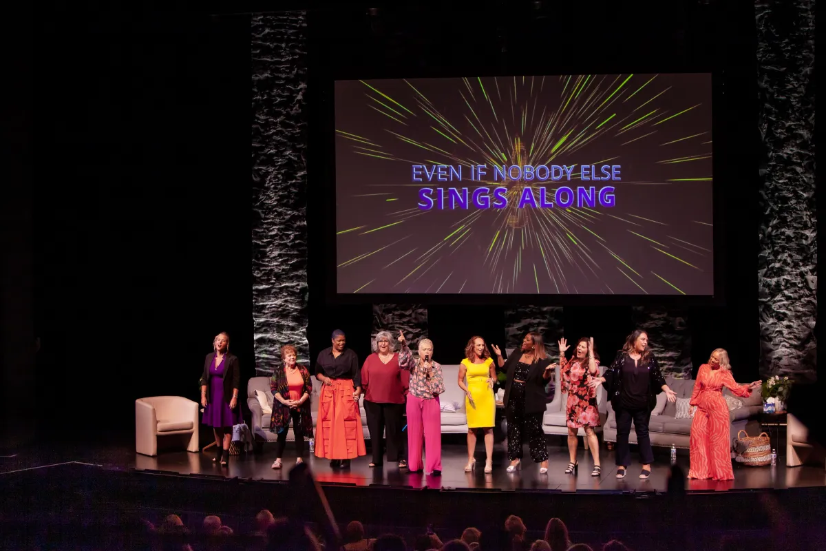 Image of a diverse group of women singing and dancing on stage with an audience in front of them