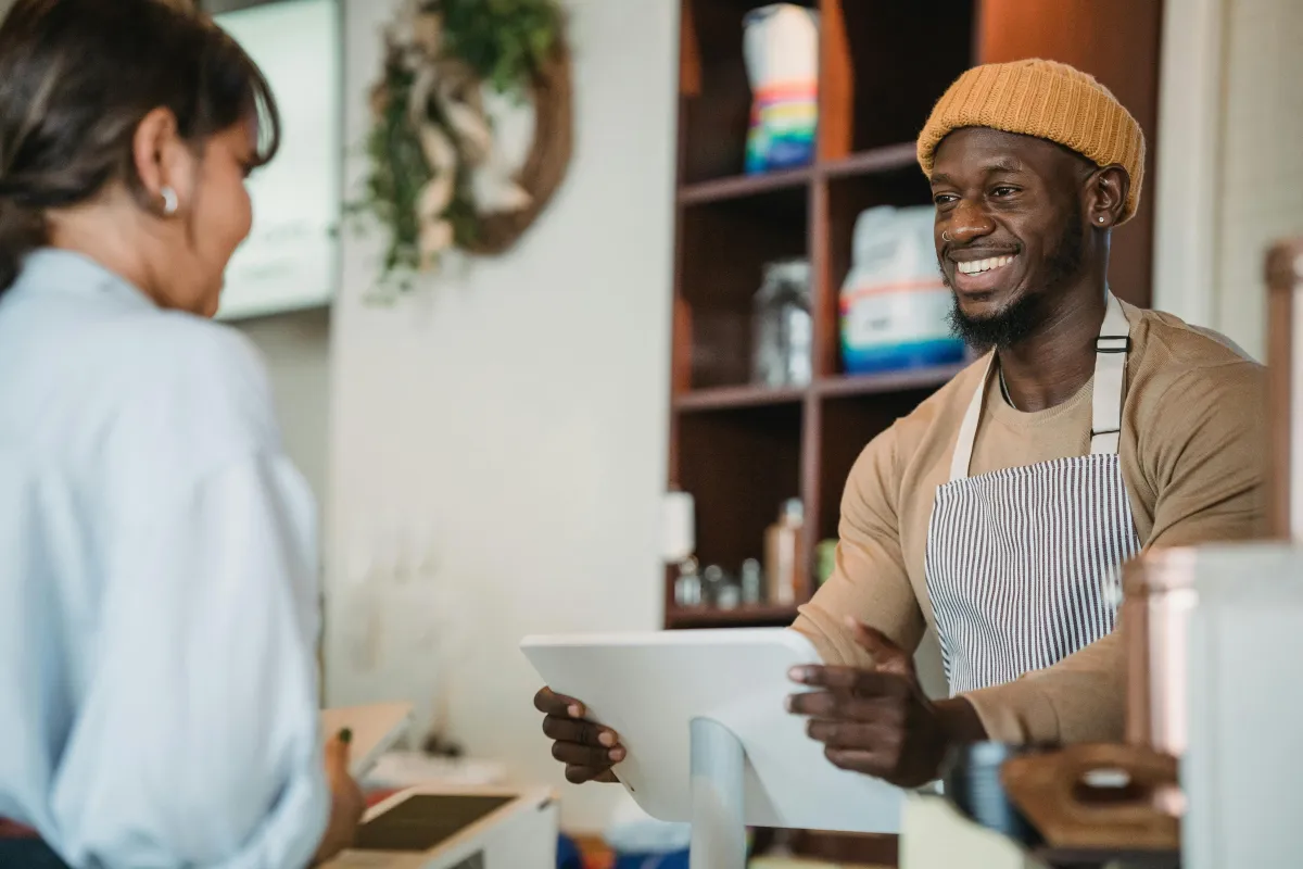 Friendly barista wearing an apron, chatting with a customer inside a cosy shop, highlighting personalised service.