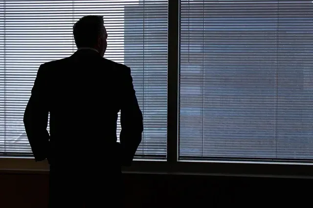 Silhouette of a businessman standing by a large window with blinds, overlooking a city skyline, symbolizing professionalism and financial expertise