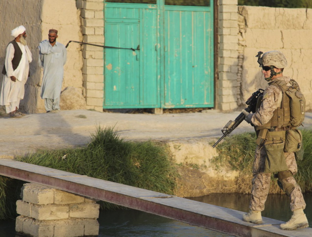 Two Afghan men in traditional garb in upper left corner. US Marine in battle dress walking across steel footbridge in Na'Wa, Helmand Province, Afghanistan.