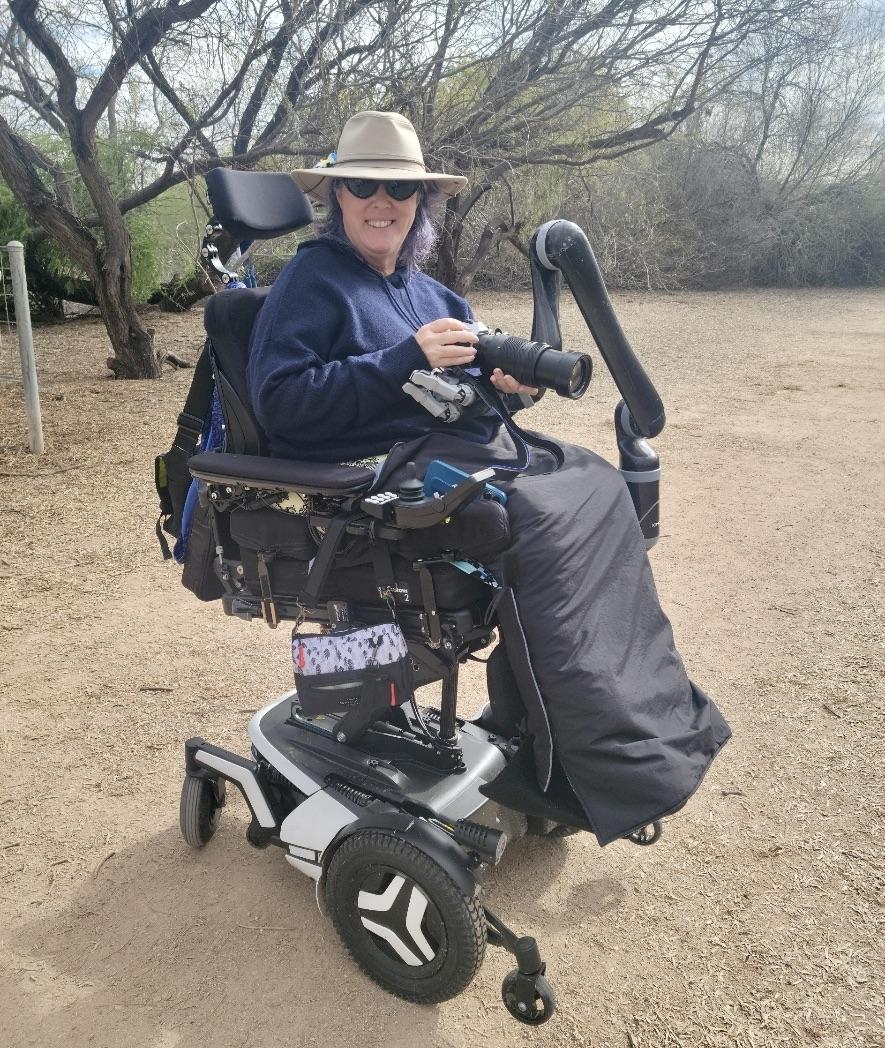 Heather sitting on her power wheelchair bundled in a sweater and lap parka wearing sunglasses and a hat holding her camera supported by a robotic arm