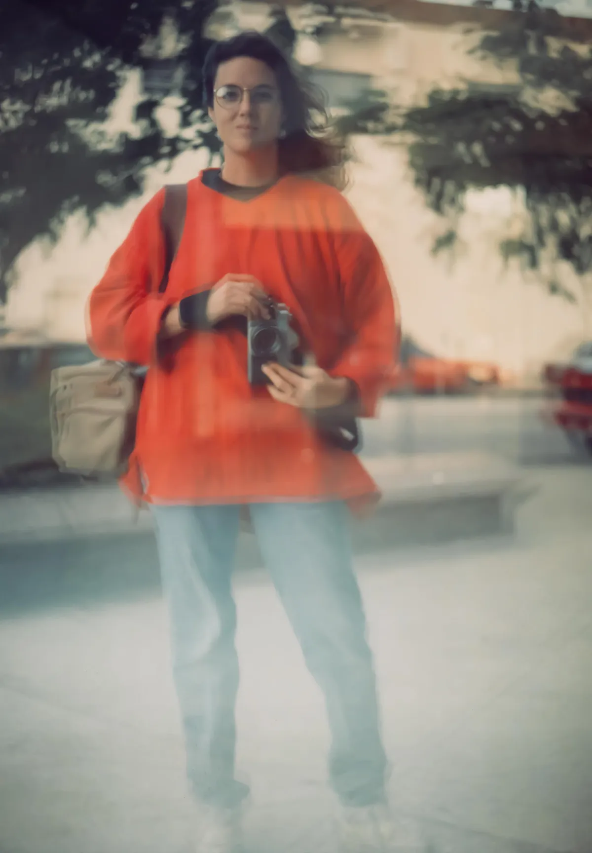 young woman with dark shoulder-length hair and glasses wearing a red cover shirt and jeans holding a camera waist high taking a selfie in a reflective building surface