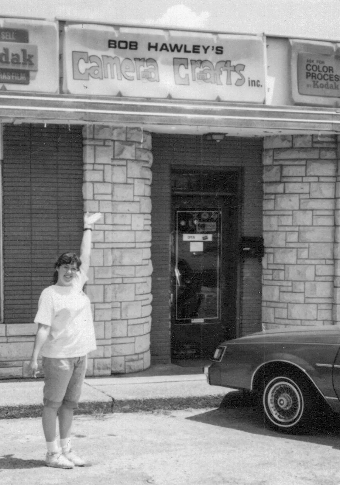 Heather in t-shirt and jean shorts holding one hand up to show the sign on the building she is smiling at the camera