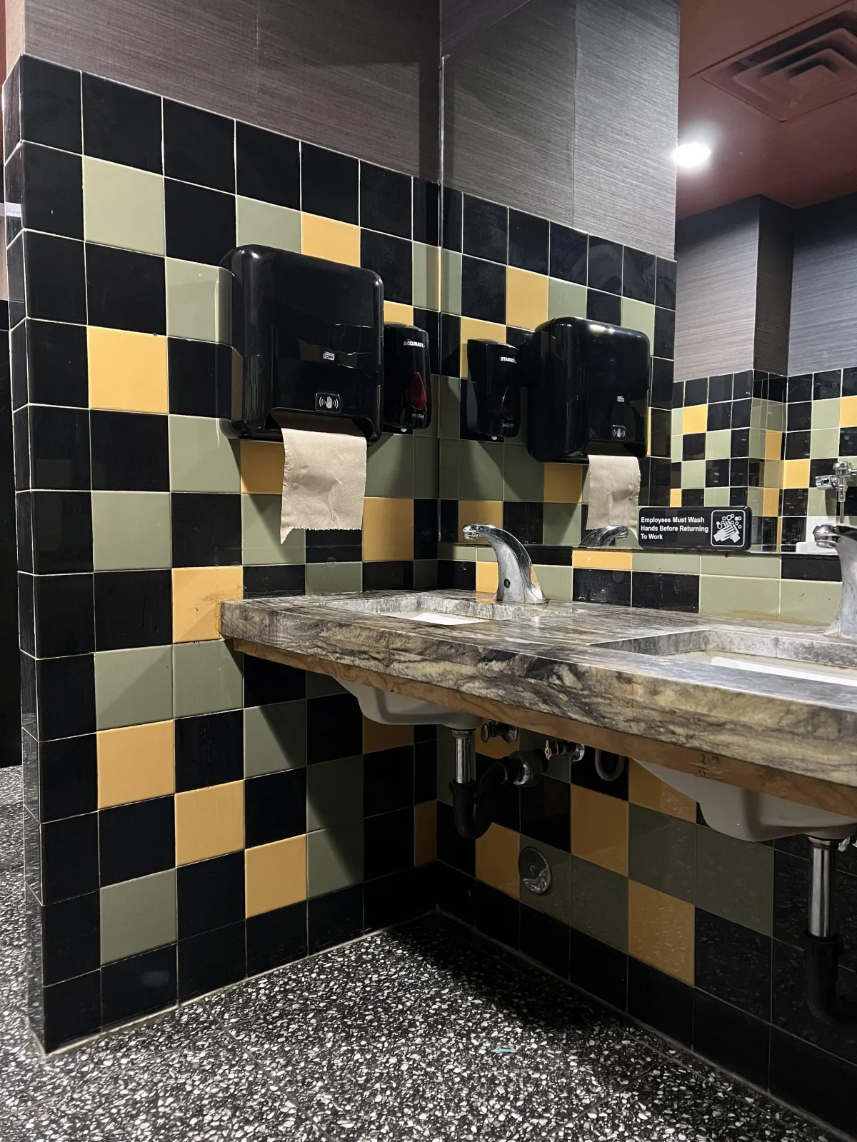 black green and yellow tiled public bathroom with soap dispenser on the mirror-side of the paper towel dispenser