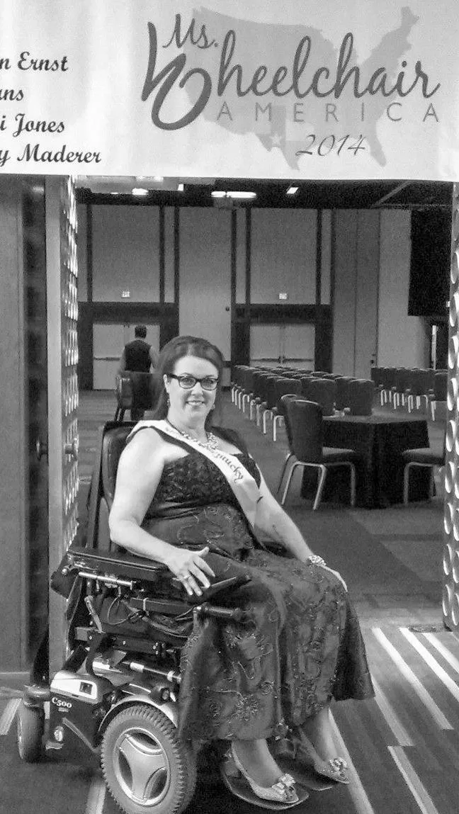 Heather sitting on her power wheelchair in formal dress and fancy dress shoes wearing a pageant sash in front of a banner for Ms Wheelchair America 2014