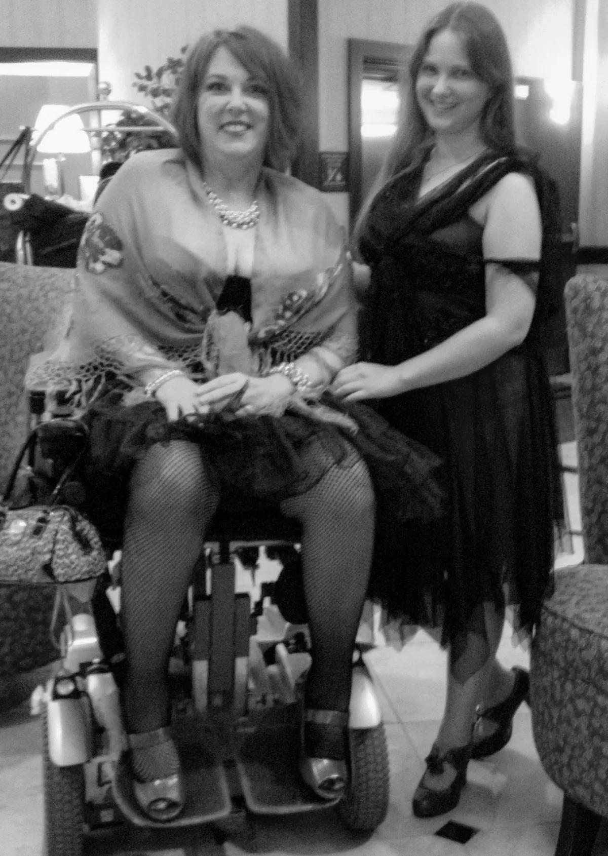 Heather looking directly at the camera while sitting on her power wheelchair she is dressed up for a fancy event wearing fishnet tights, high heeled shoes a ruffled skirt a corset and a shawl her friend is standing next to her