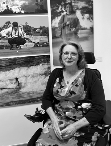 Heather sitting on a power wheelchair in a short fancy dress holding a glass of champagne in front of photos on a gallery wall