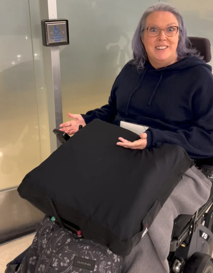 Heather sitting on her power wheelchair with a cushion on her lap next to an automatic door button - she looks exasperated