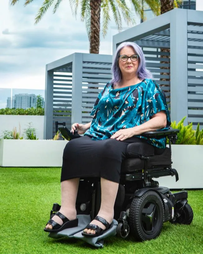 Heather sitting on her power wheelchair smiling at the camera wearing a teal and black top black skirt and black sandals