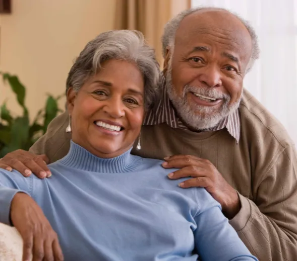 A senior couple standing together in a lush green park, both smiling warmly.