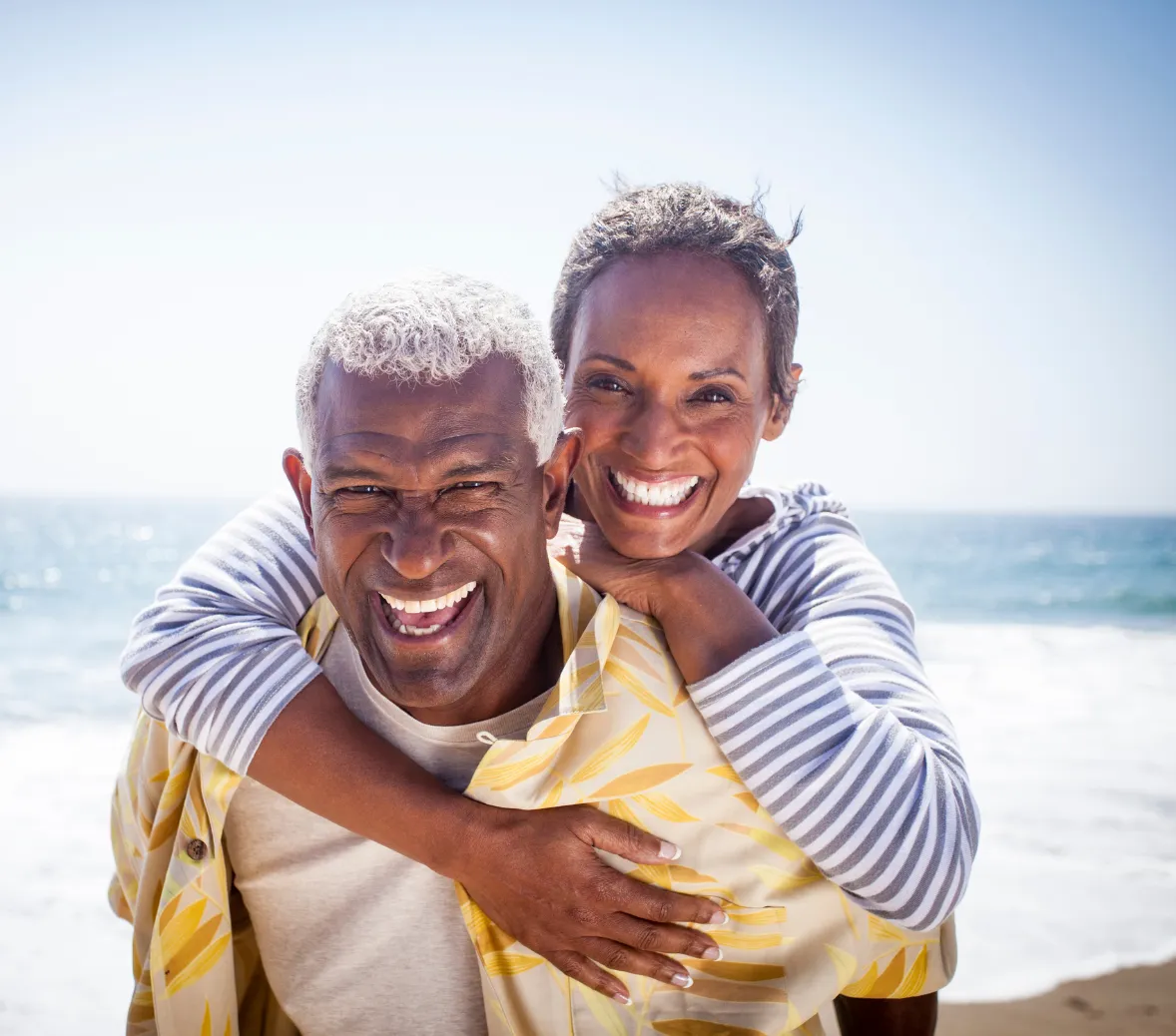 Happy senior couple smiling outdoors; woman in a yellow sweater embraces man with glasses and a gray shirt from behind.