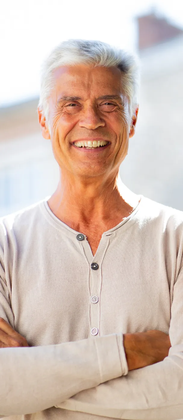 Smiling older man with silver hair wearing a beige henley shirt, standing confidently outdoors with crossed arms.