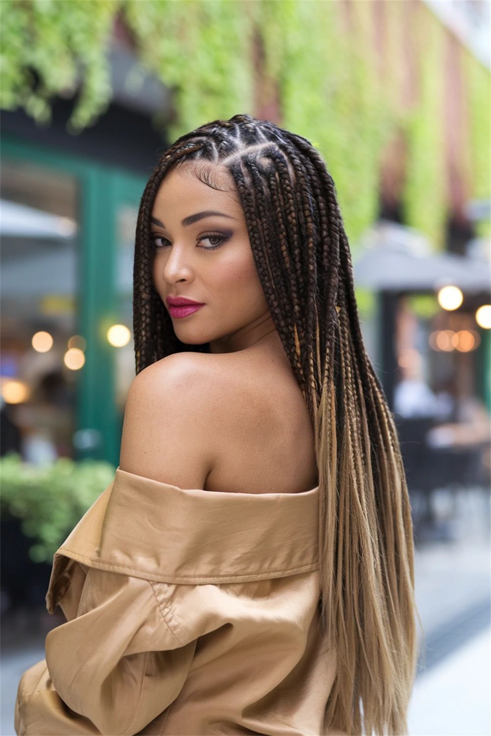Portrait of a woman with long braided hair, wearing a beige off-the-shoulder top, looking over her shoulder.