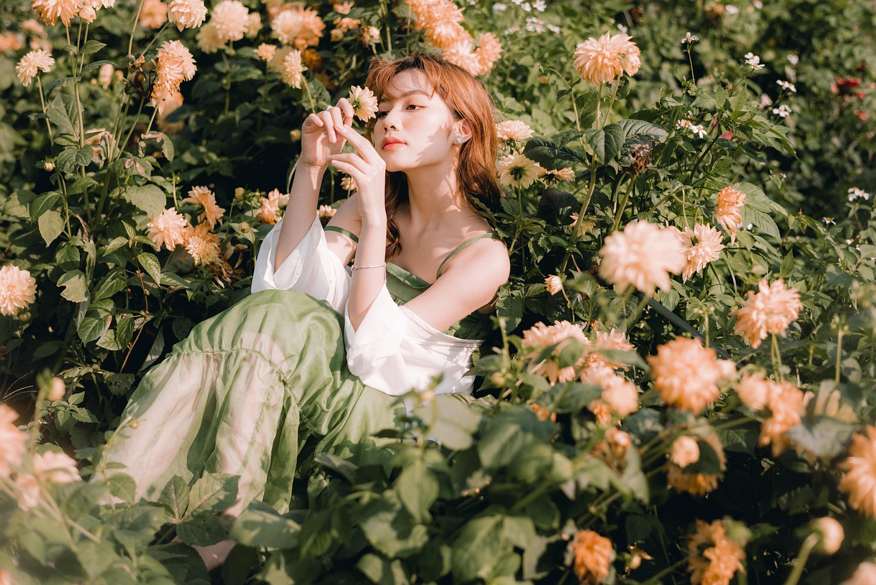 women slightly smiling sitting in field of pink/coral flowers, she is looking at one of the flowers that she has in her hands