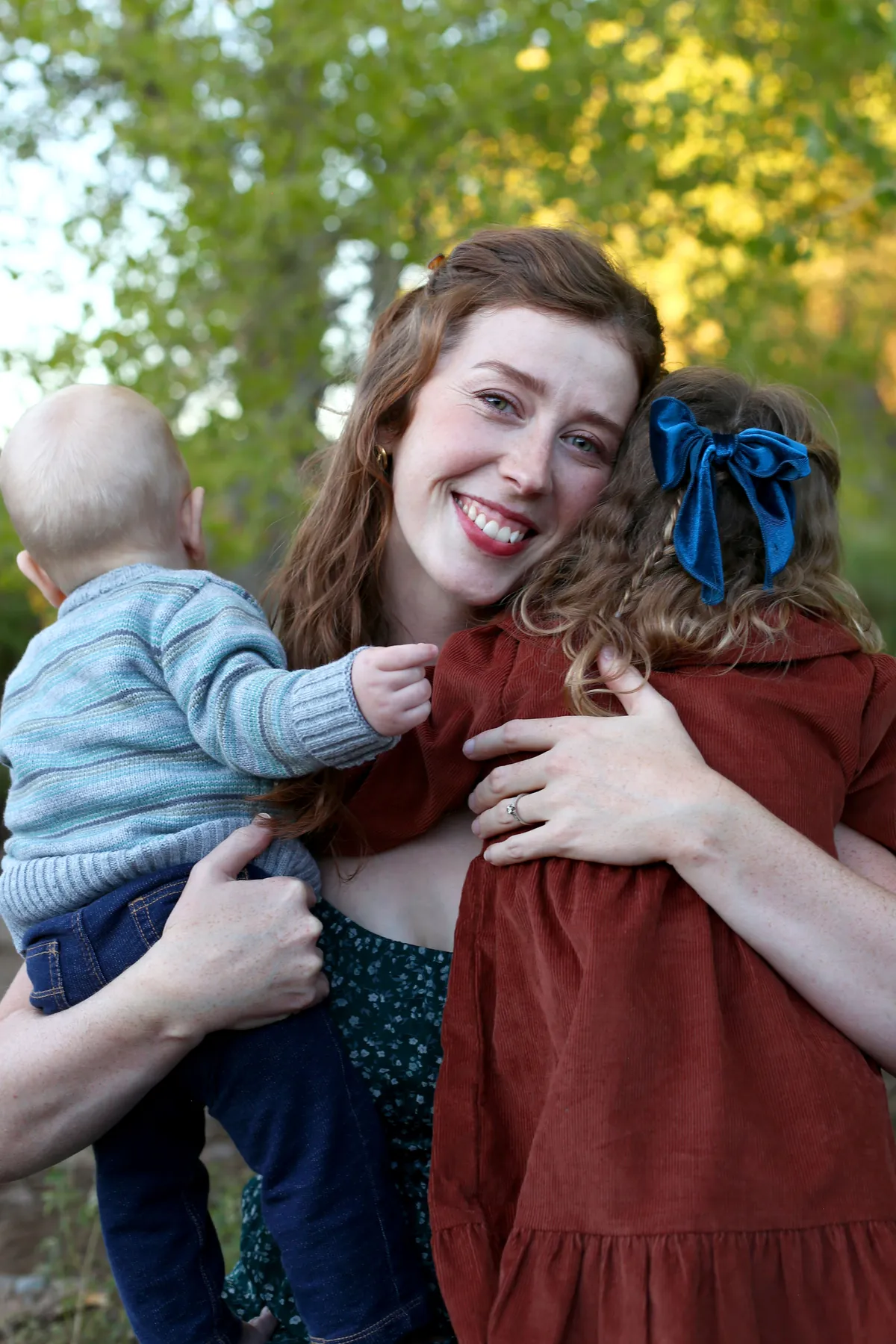 Hannah of Know and Nurture hugging her young daughter and baby boy, outdoors with green trees in the background.