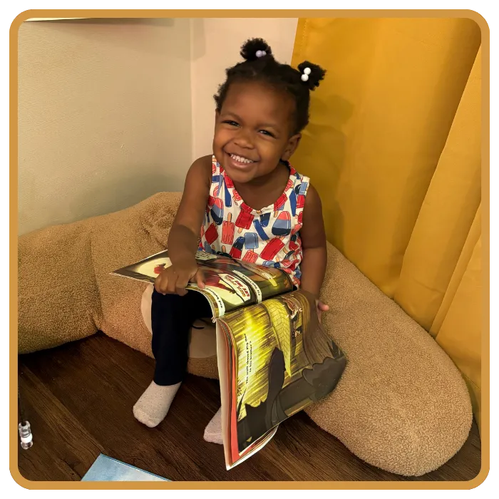 A smiling toddler with two puffs in her hair sits on a large plush couch holding an open book with colorful illustrations in Smyrna, GA