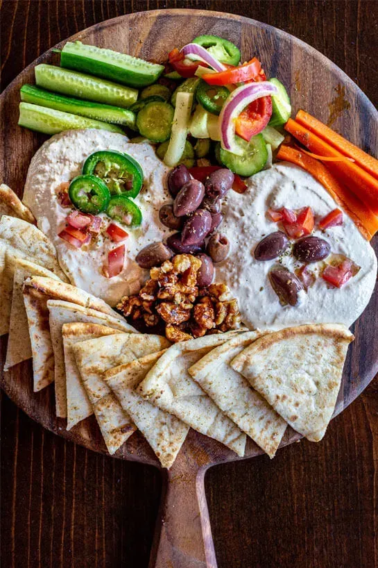 Wooden serving board featuring hummus topped with olives, walnuts, and jalapeños alongside fresh veggies and pita bread from Houston’s stress-free catering solution.