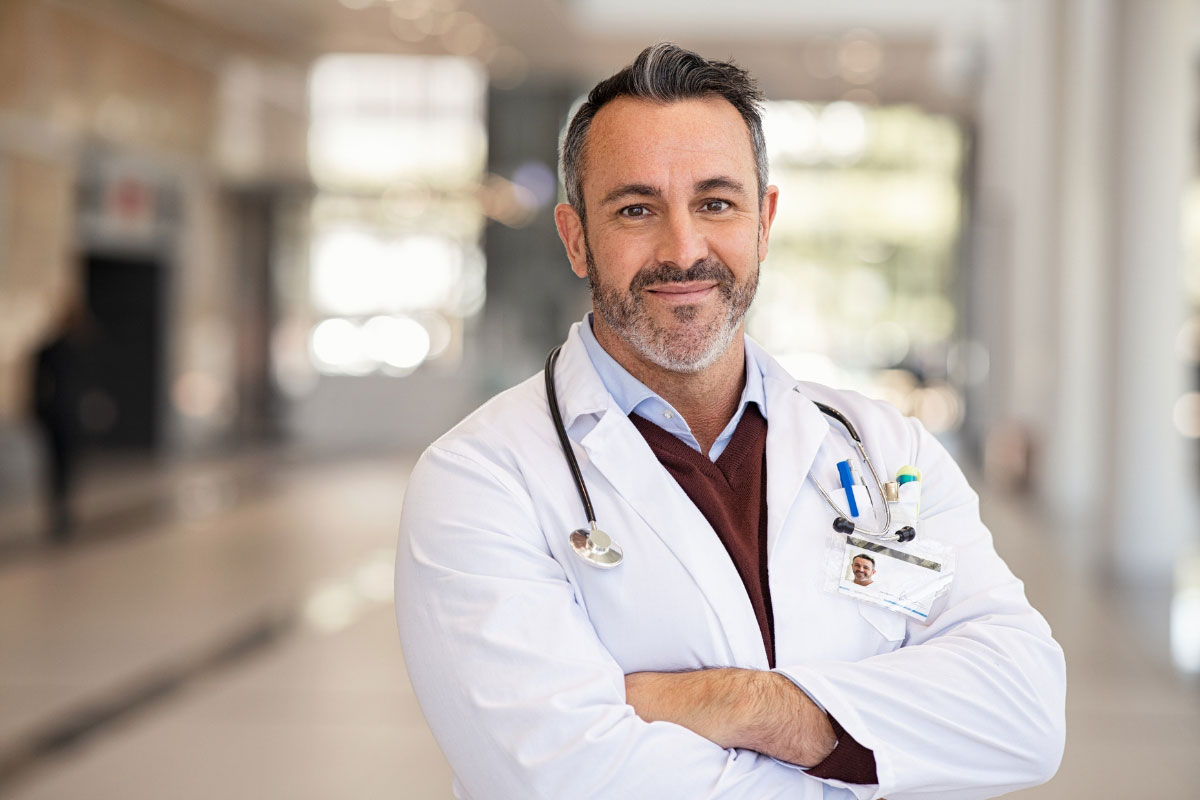 Portrait of a confident, smiling male physician in a white lab coat with crossed arms and a stethoscope around his neck in a modern hospital lobby.