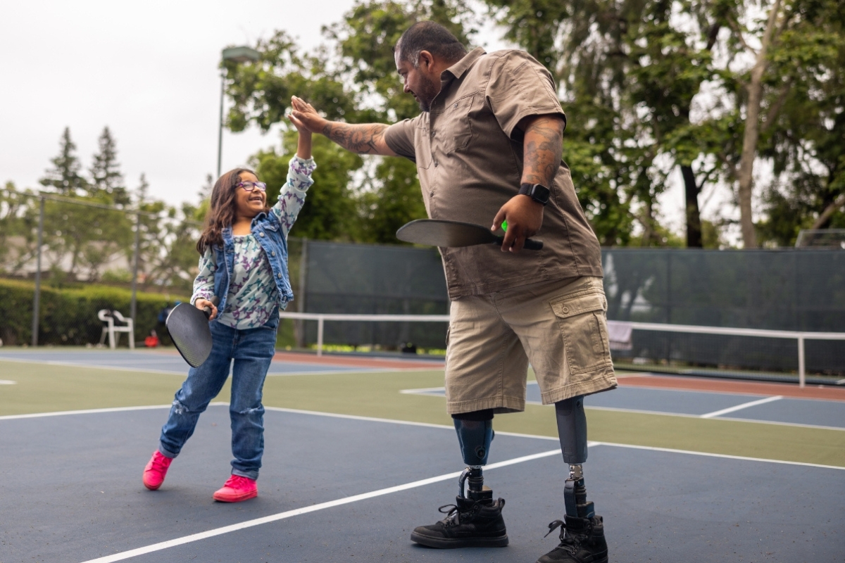 Parent with bilateral leg prostheses high-fiving a child during an outdoor activity