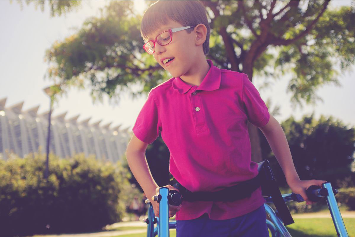 Young boy wearing glasses and a pink polo shirt using a blue gait trainer to walk outside in a park