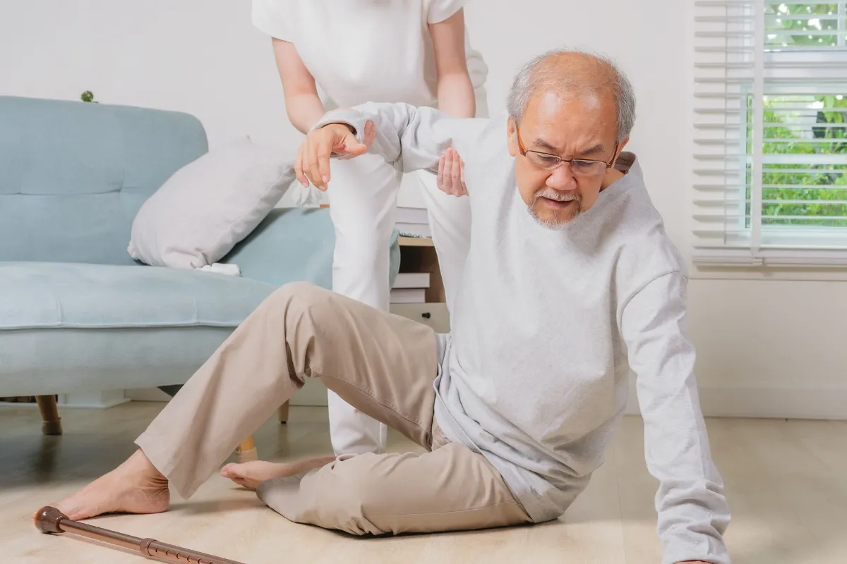 Caregiver assisting an older adult who has fallen on the floor near his walking cane