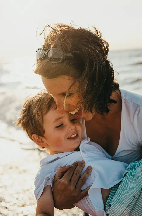 Smiling woman holding and cuddling her young son at the beach during sunset, both wearing white tops and bathed in warm golden light, capturing a joyful and tender moment of motherhood and connection.