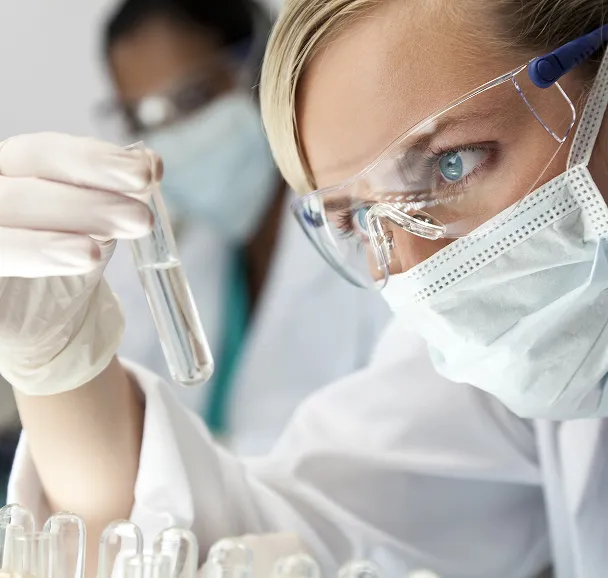 Female scientist wearing safety goggles, a face mask, and gloves, examining a test tube in a laboratory setting.