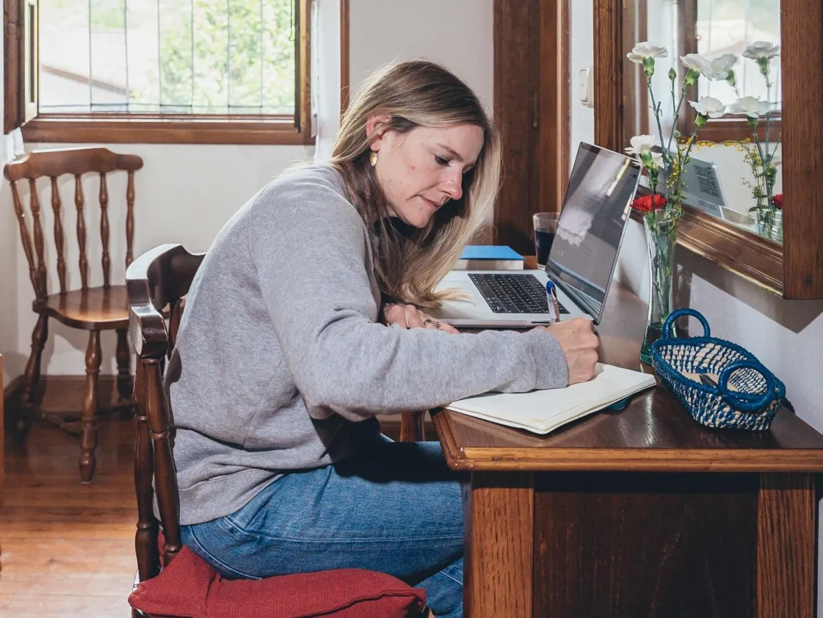 Woman sitting at a desk writing in a notebook next to a laptop in a cozy room at a retreat
