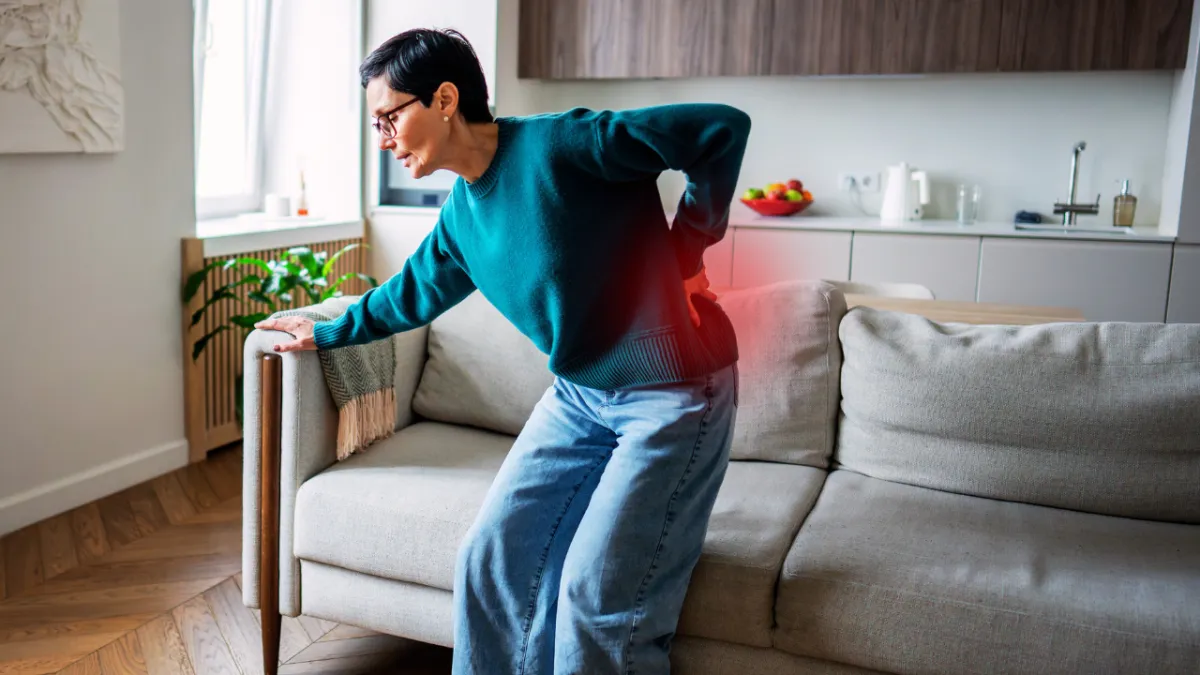 A woman in a living room clutching her lower back in severe pain with a red glow overlay on her lumbar spine, showcasing bulging disc symptoms treated in Fort Mill, SC.