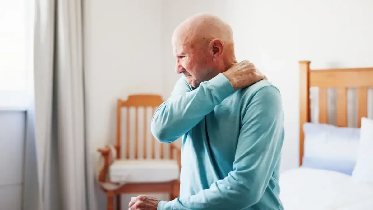 An elderly man sitting on his bed clutching his painful shoulder in distress, illustrating severe stiffness and age-related frozen shoulder symptoms treated in Fort Mill, SC.
