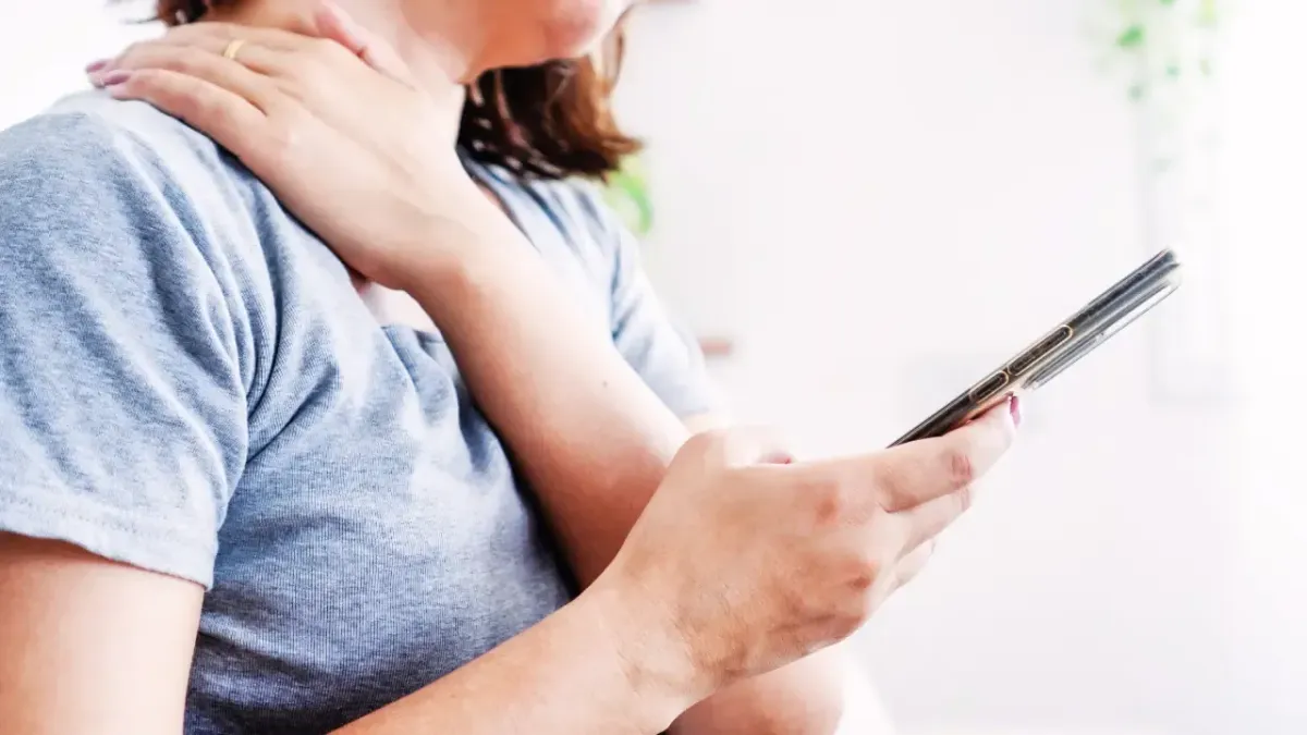 A woman holding a smartphone while massaging her sore neck and shoulder muscles, illustrating text neck symptoms treated with chiropractic care in Fort Mill, SC.