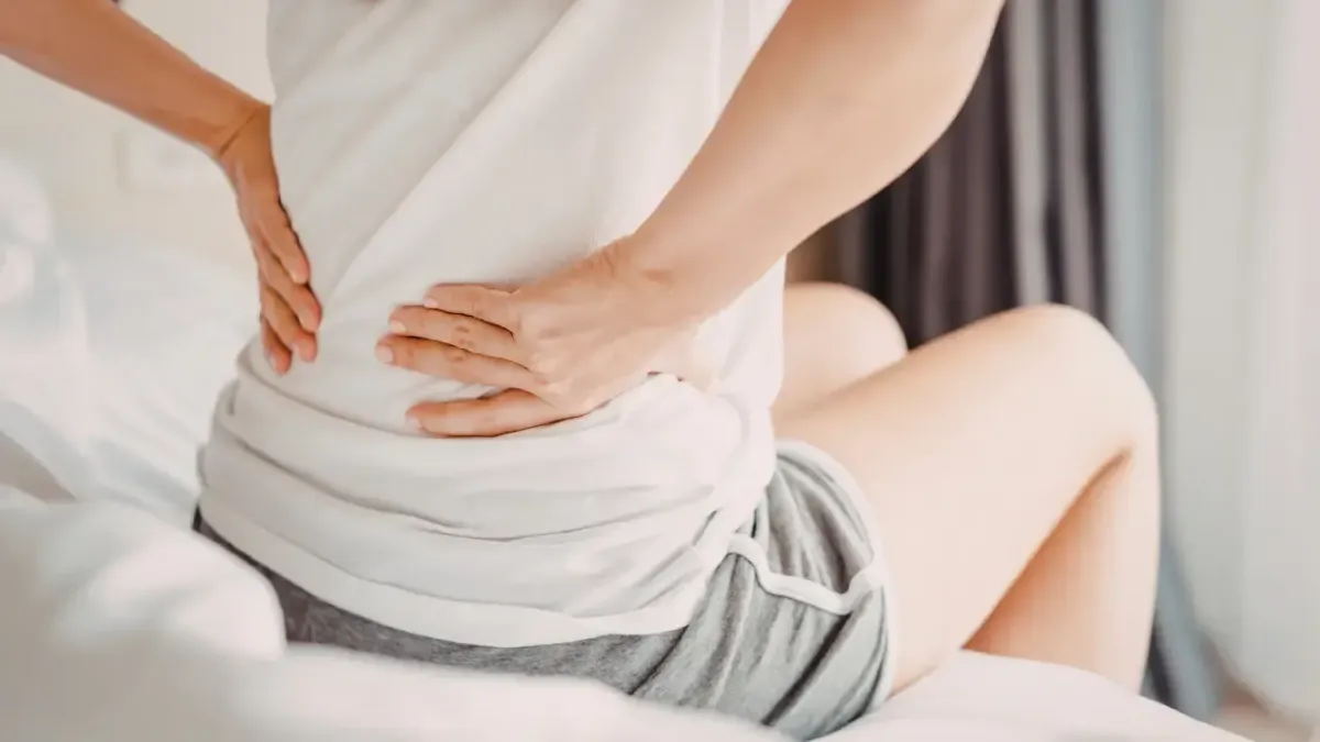 A woman sitting on the edge of a bed clutching her painful lower back in the morning, illustrating arthritic symptoms managed with degenerative joint disease treatment in Fort Mill, SC.